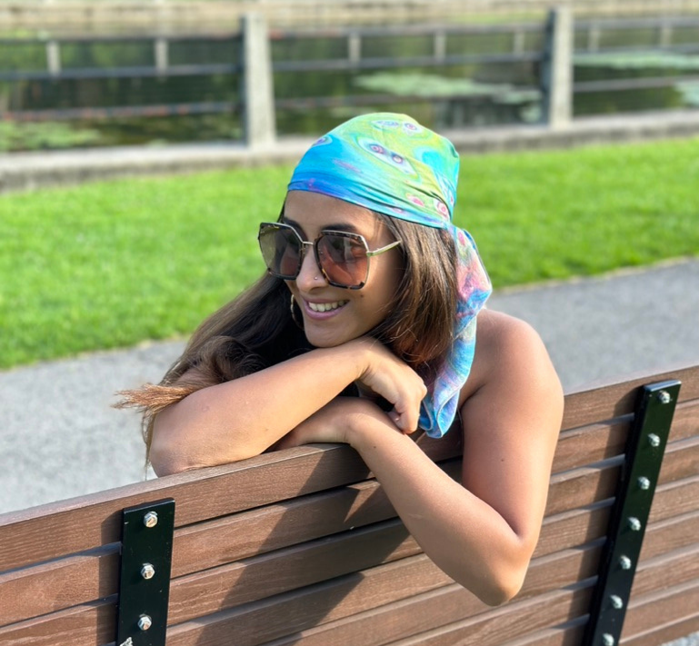 Luxury Silk Scarf Woman wearing a colorful headscarf and sunglasses, sitting on a bench outdoors.